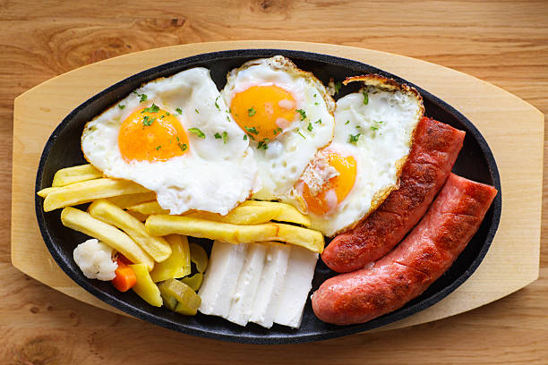 Breakfast with sunny side up eggs, sausage, cheese, French fries, pickled vegetables served on black oval plate. Directly Above.